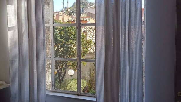 A room with a large window, white curtains, and a view of buildings and trees outside.