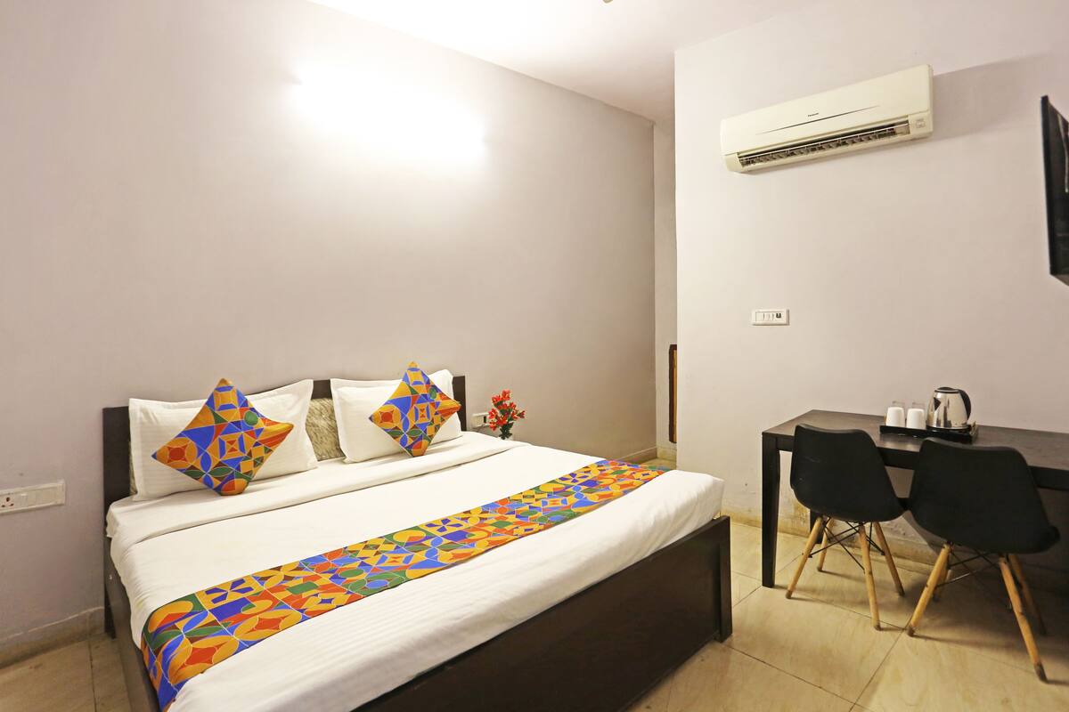 A hotel room with a bed, a desk, two chairs, an air conditioner, and a small table with a kettle.