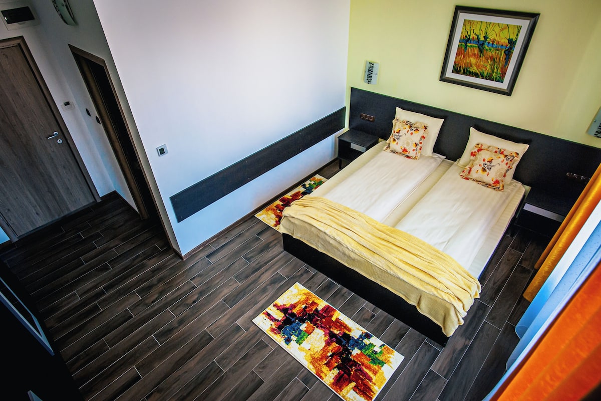 A bedroom with a large bed, a small bedside table, a colorful rug, and a painting on the wall.