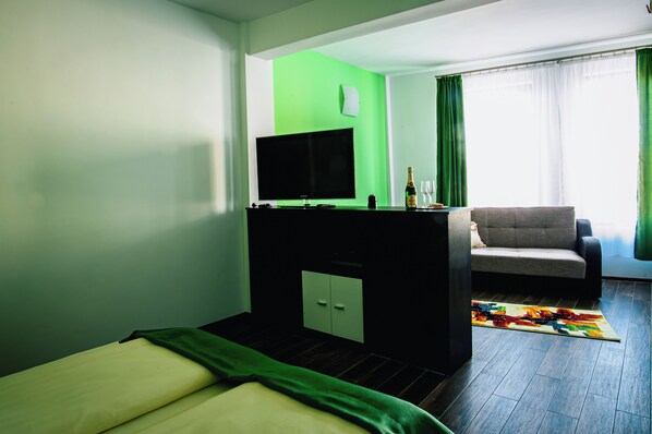 Habitación de hotel con cama, televisión, escritorio con una botella y vasos, sofá y una alfombra colorida.