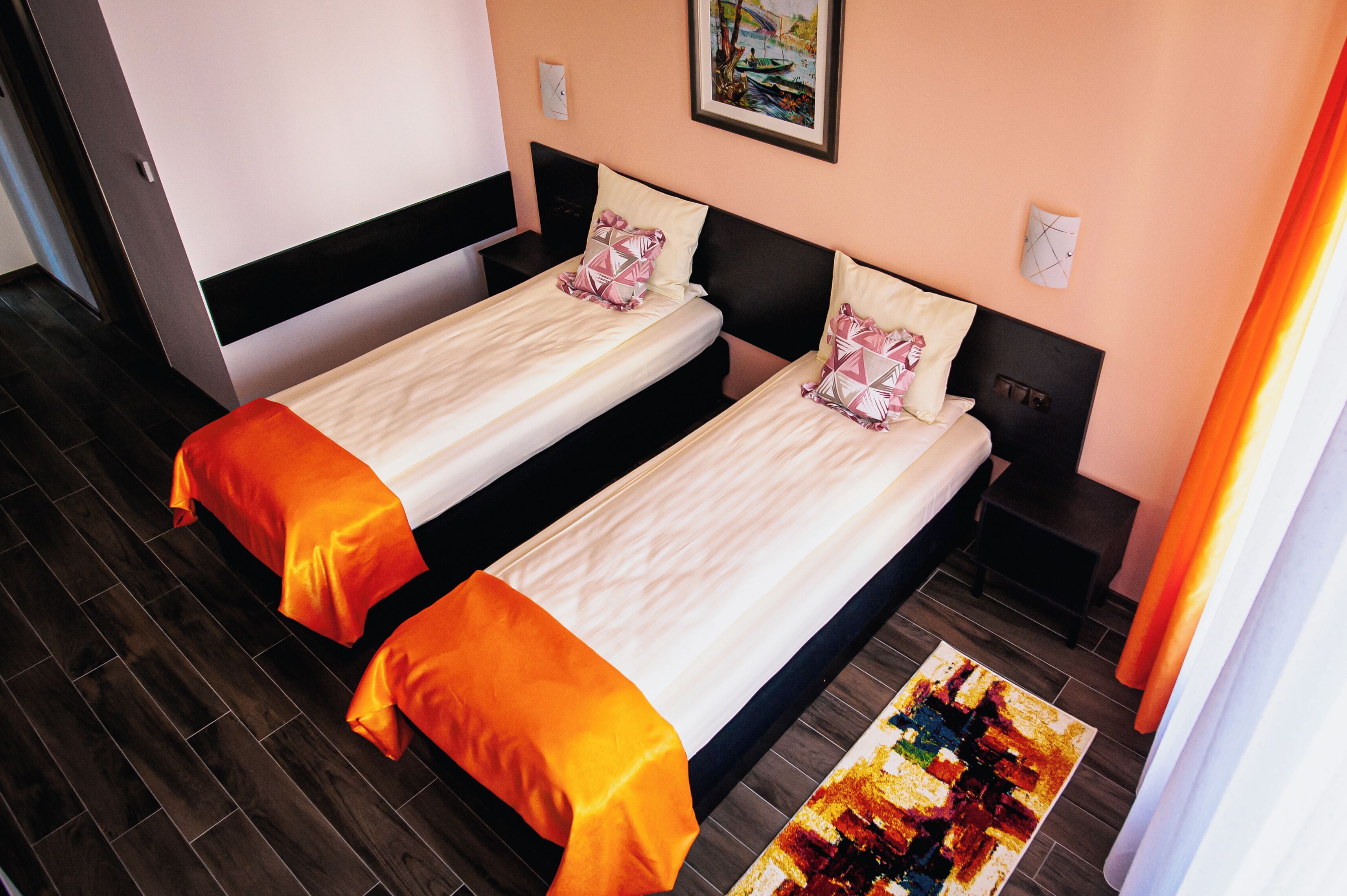 A hotel room with two single beds, a wooden floor, a painting on the wall, and a window with curtains.