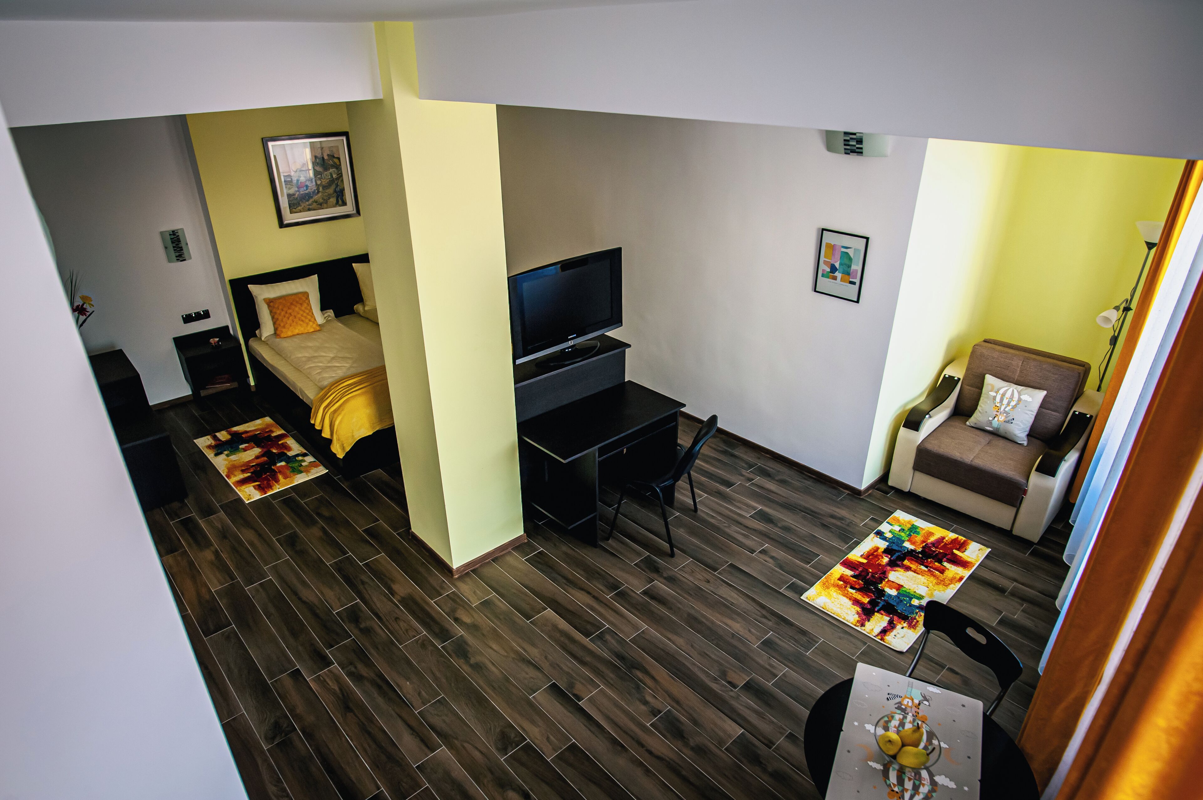 A compact hotel room with a bed, a desk with a TV, a small sofa, and a dining area.