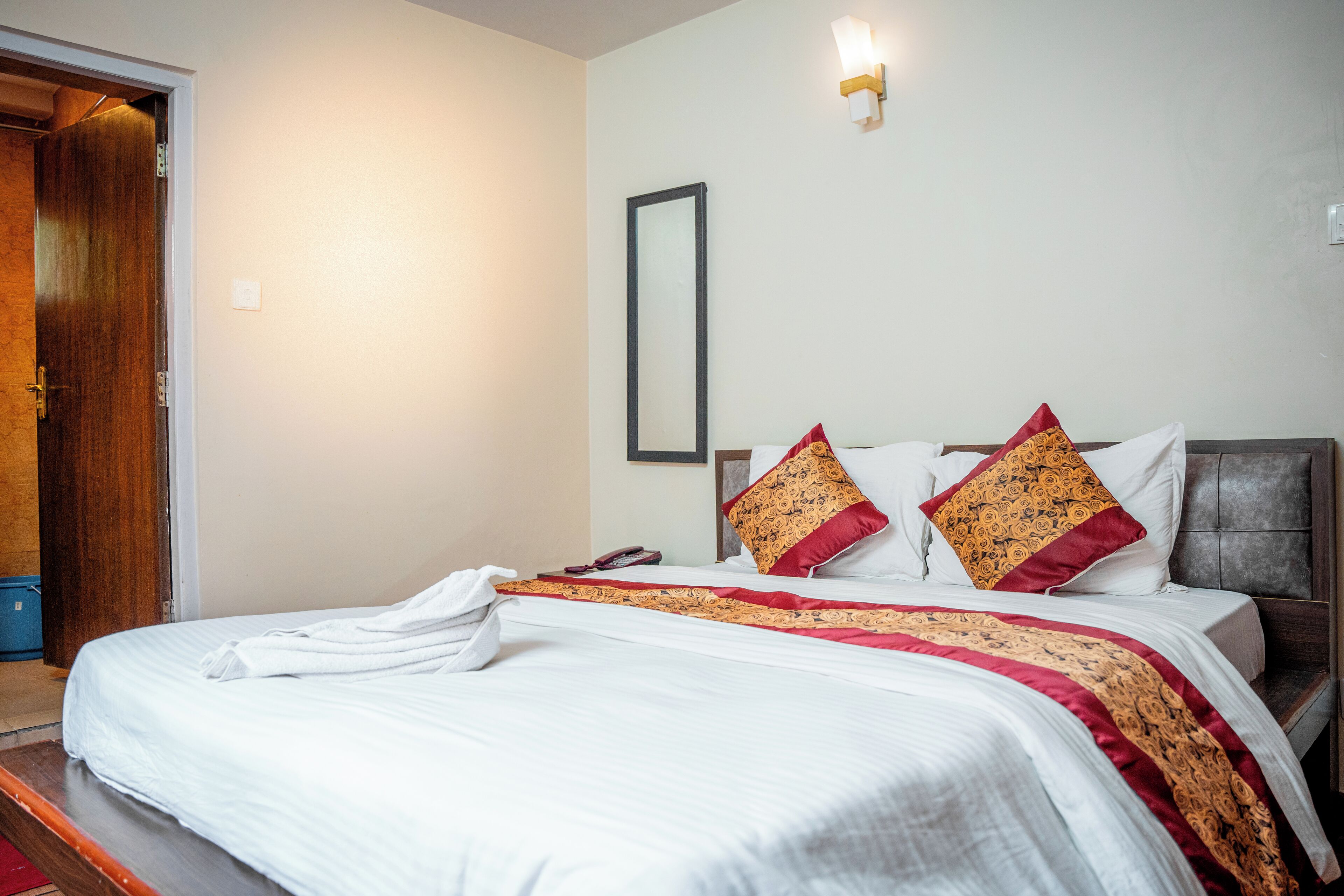 A neatly made bed with a white and red patterned bedspread, two decorative pillows, a bedside table with a lamp, and a door leading to another room.