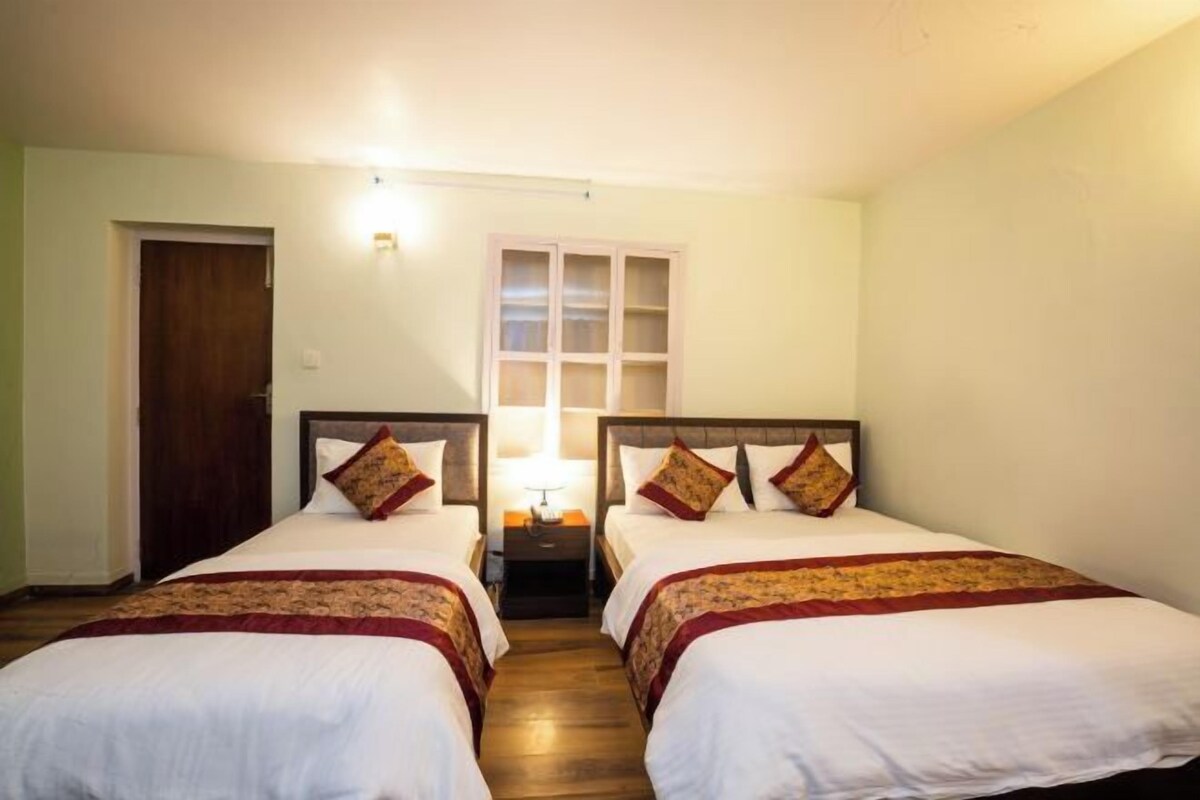 Habitación de hotel con dos camas individuales, una puerta de madera, una ventana con persianas y una mesita de noche con lámpara.