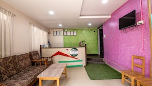 A reception area with a sofa, wooden table, and chairs, a counter with a sign, and a TV mounted on a pink wall. - Hotel Harmony Bistro (Lalitpur)