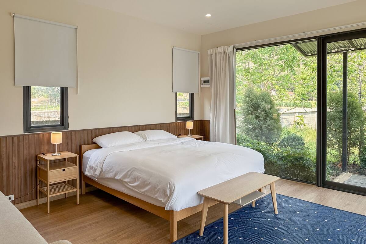A modern hotel room with a large bed, a bedside table with a lamp, a bench, and a view of greenery through large windows.