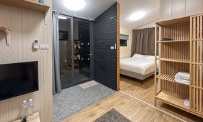 Quarto de hotel moderno com porta de vidro deslizante, cama, televisão e guarda-roupa de madeira.