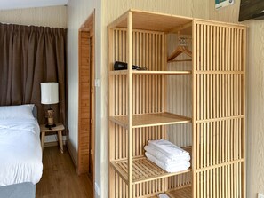 Quarto de hotel com uma cama, uma mesa de cabeceira com abajur e um guarda-roupa de madeira com prateleiras.