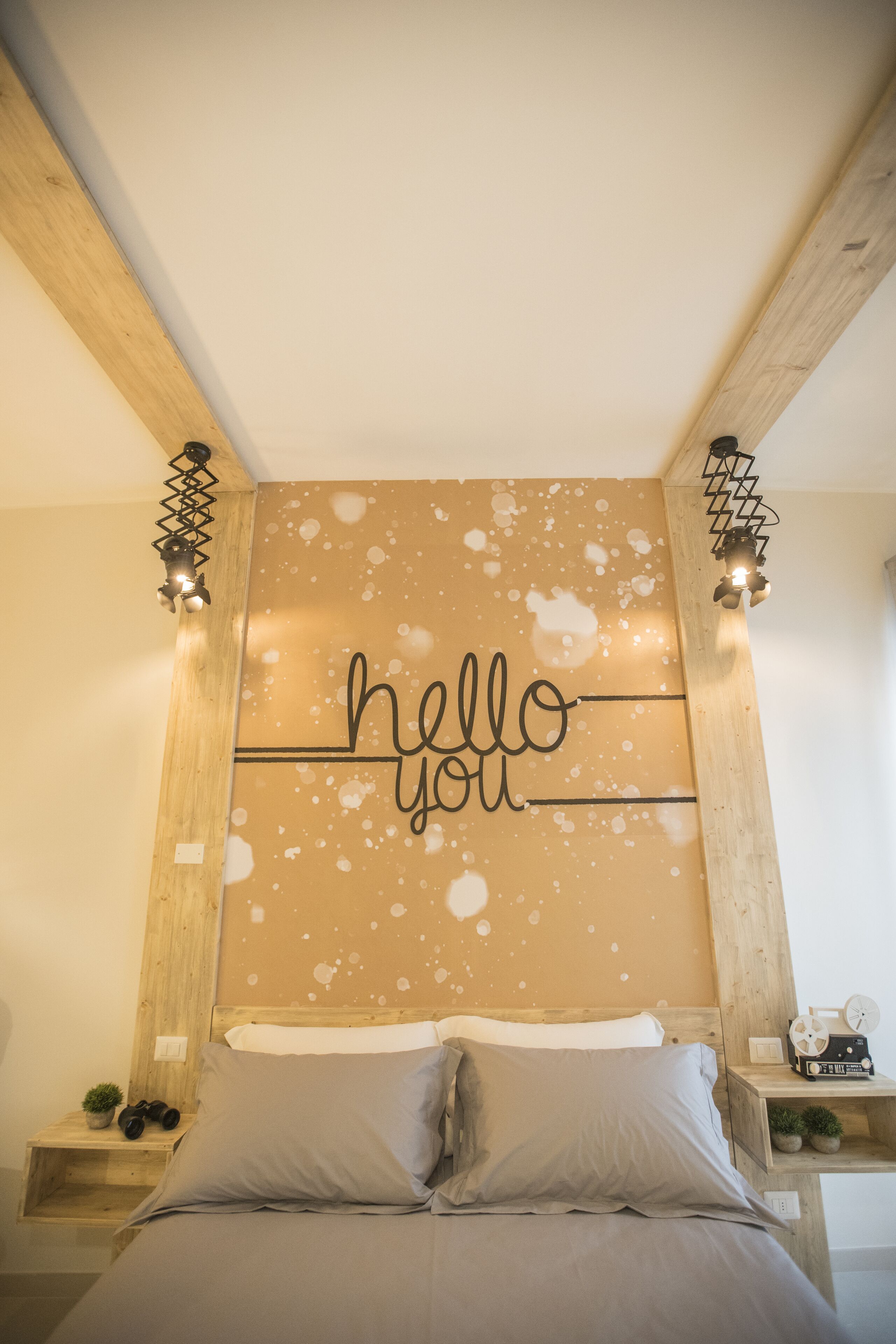 A bedroom with a wooden headboard, a bed with grey bedding, a wall with a 'hello you' sign, and two bedside tables with small plants and a clock.