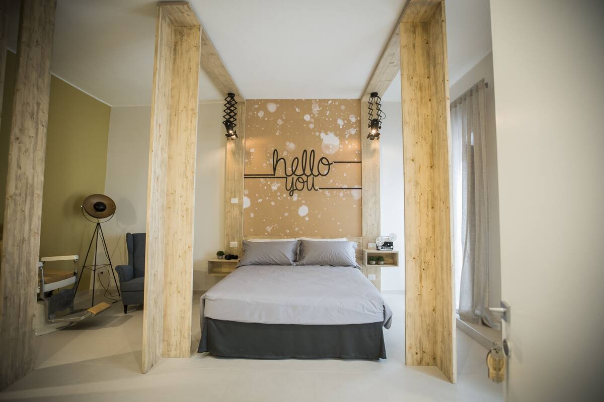 Et soveværelse med en seng, et skilt med teksten 'hello you', træbjælker og et lille bord med en lampe.