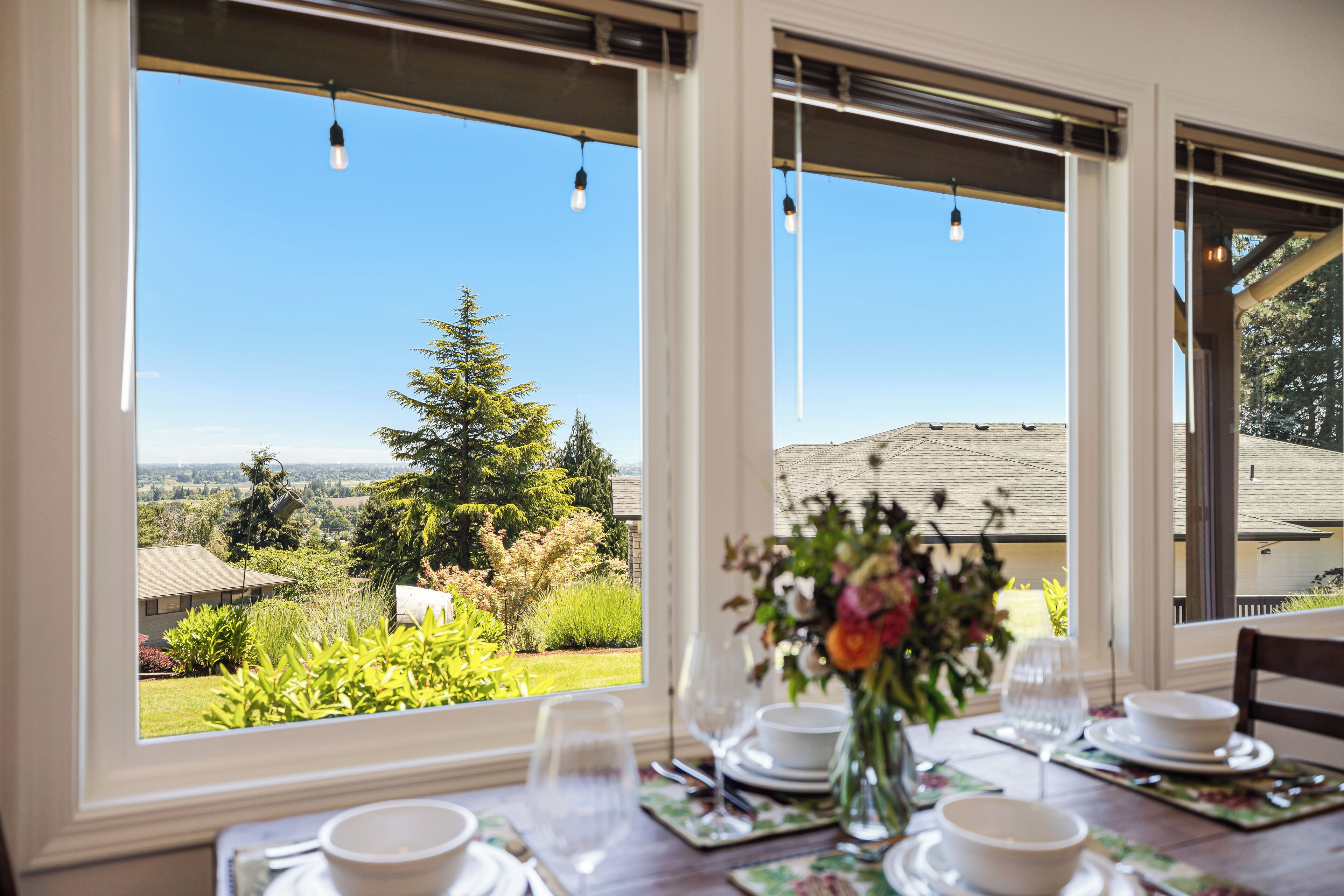 A dining table set with white plates, bowls, and glasses, with a vase of flowers in the center. Large windows provide a view of a garden and a distant building.