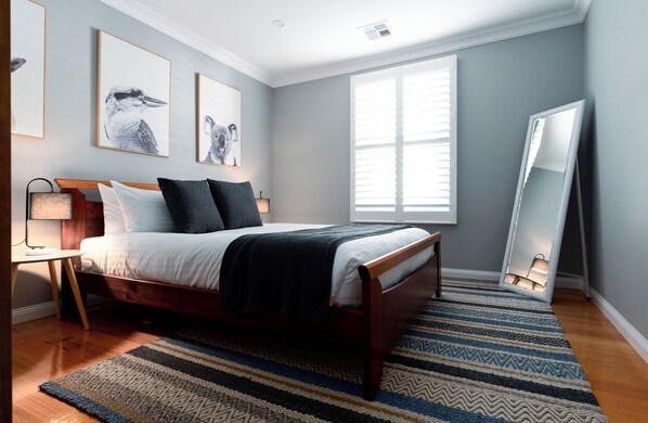 A bedroom with a wooden bed, a nightstand, a lamp, a large window with blinds, and a mirror leaning against the wall.