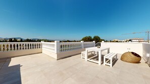 Eine Dachterrasse mit weißem Tisch und Stühlen, einem gewebten Korb und Blick auf die Stadtlandschaft.