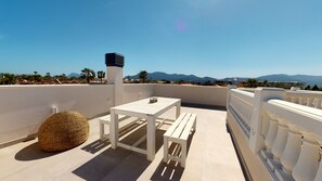 Une terrasse sur le toit avec une table et des bancs blancs, un panier en osier, et une vue sur les montagnes et les palmiers.