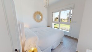 Un dormitorio con cama, lámpara de noche, espejo y ventana con vistas a la vegetación.