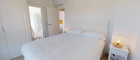 Een witte slaapkamer met een groot bed, een nachtkastje met een lamp en een airconditioner boven het bed.