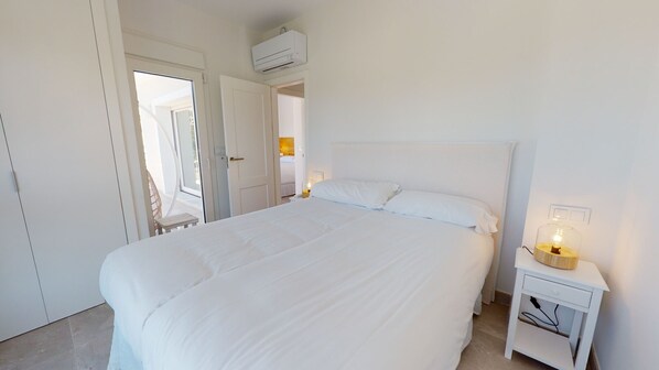 A white bedroom with a large bed, a bedside table with a lamp, and an air conditioning unit above the bed.