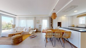 A modern living room with a sofa, dining table, and chairs, a kitchen with a large island, and a view of the outdoors. - Greenside oasis villa by ON Travel (Oliva)