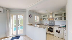 A modern kitchen with a built-in oven, microwave, and washing machine, a large window with a view of a pool, and a clock on the wall.