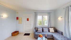 A living room with a sofa, a window with curtains, a door, and a wall-mounted light fixture.