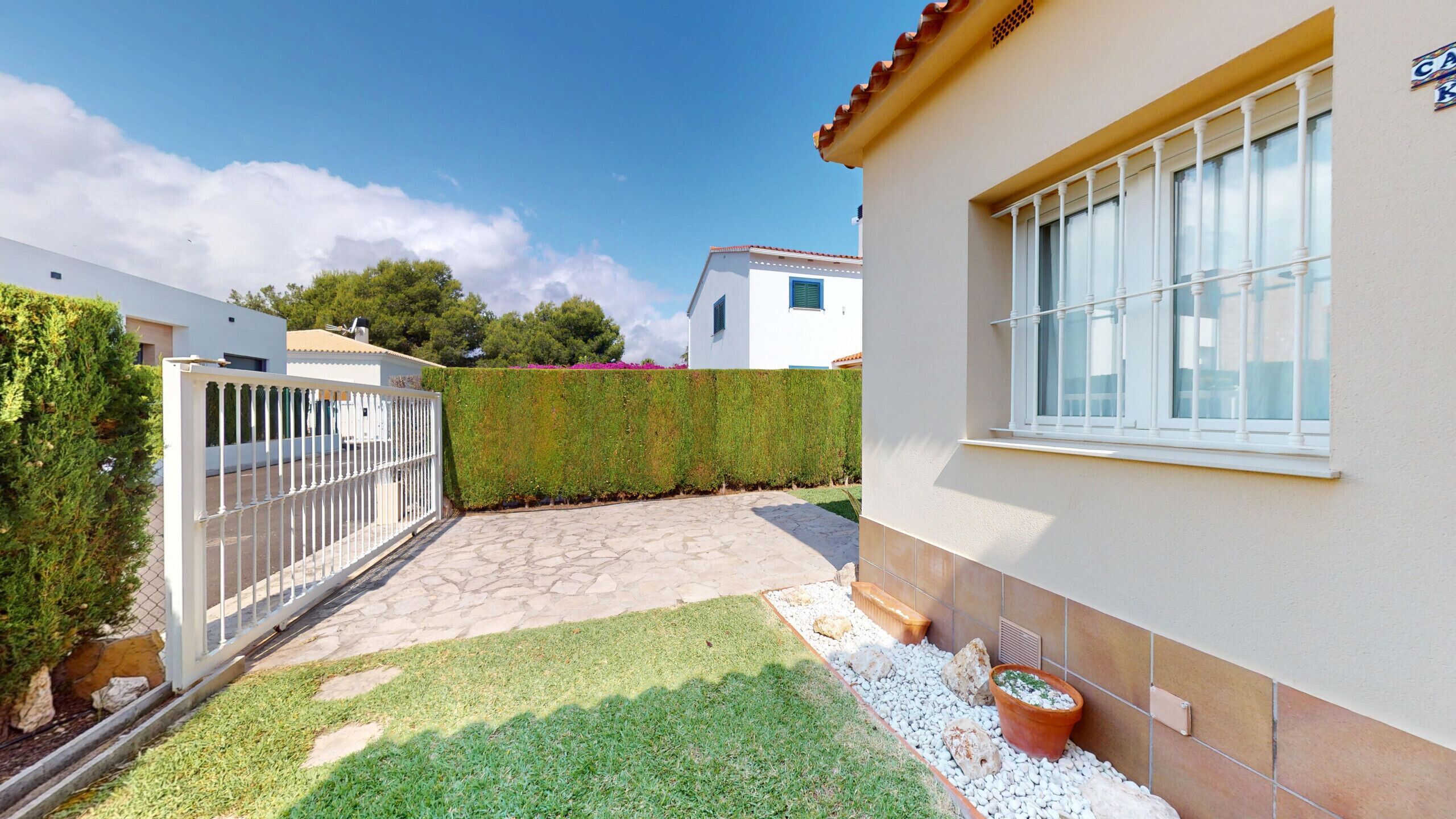 A residential property with a paved patio, a white fence, a neatly trimmed hedge, and a window with a white balustrade.