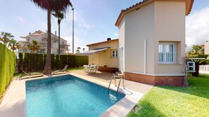 A swimming pool with a sun lounger, a table with chairs, and a covered patio area.