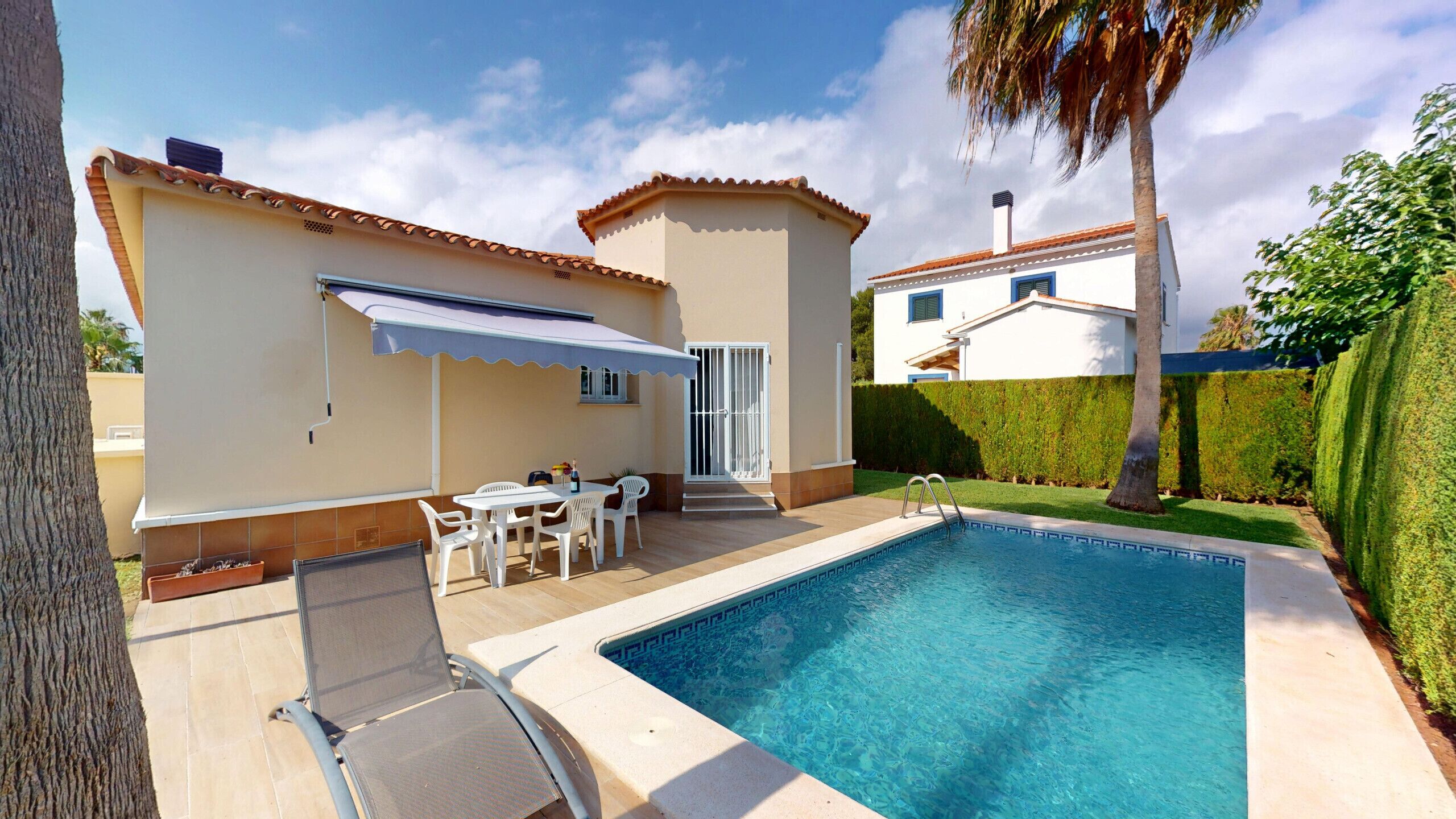 Ein Poolbereich mit überdachter Terrasse, einem Schwimmbecken und einem zweigeschossigen Haus mit weißer Fassade und blauen Fensterläden.