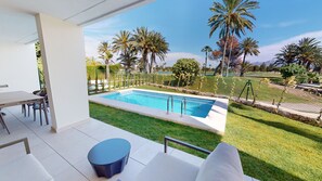 Zona de piscina amb taula i cadires, una taula rodona petita i vistes a una piscina envoltada de palmeres i un prat.