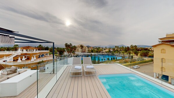 A rooftop pool area with lounge chairs, a view of a swimming pool, and surrounding buildings.
