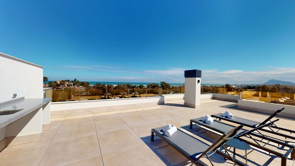 A rooftop terrace with a clear view of the ocean and mountains, furnished with lounge chairs and a small table.