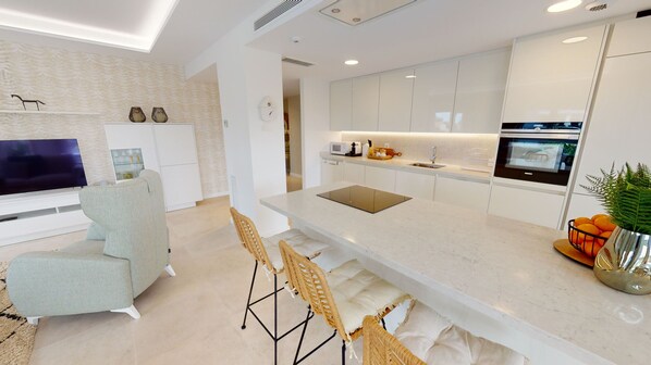 A modern kitchen with white cabinetry, a built-in oven, and a dining area with a light-colored sofa and wicker chairs.