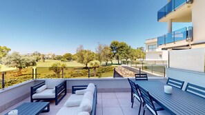 A balcony with outdoor seating, a view of a golf course, and a modern building with glass balconies.