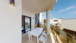Un balcon avec une table et des chaises blanches, une porte vitrée donnant sur un espace intérieur, et une vue sur les bâtiments voisins.