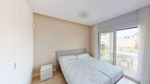 Ein Schlafzimmer mit Bett, Nachttisch, Schublade und einer Glastür, die zu einem Außenbereich mit Tisch und Stühlen führt.