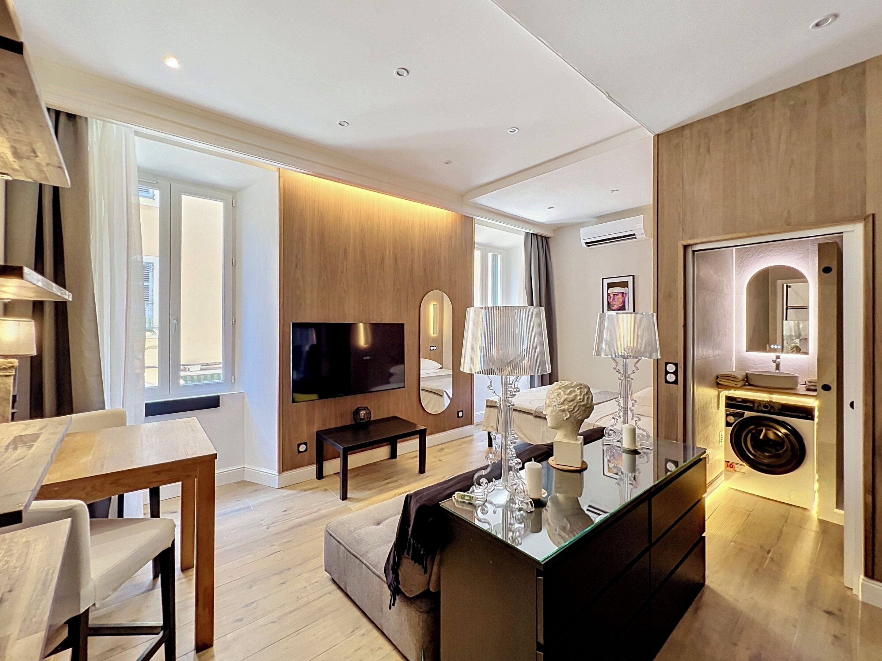 A modern hotel room with a flat-screen TV, a sofa, a dining table, a coffee table with decorative items, a mirror, and a washing machine.