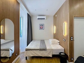 A hotel room with a bed, a mirror, an air conditioning unit, a framed picture on the wall, and a door with a light switch.