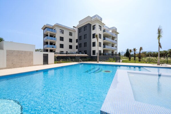 A swimming pool with a clear blue water surface, surrounded by a modern building with balconies and a flat roof. - Prímula by ON Travel (Oliva)