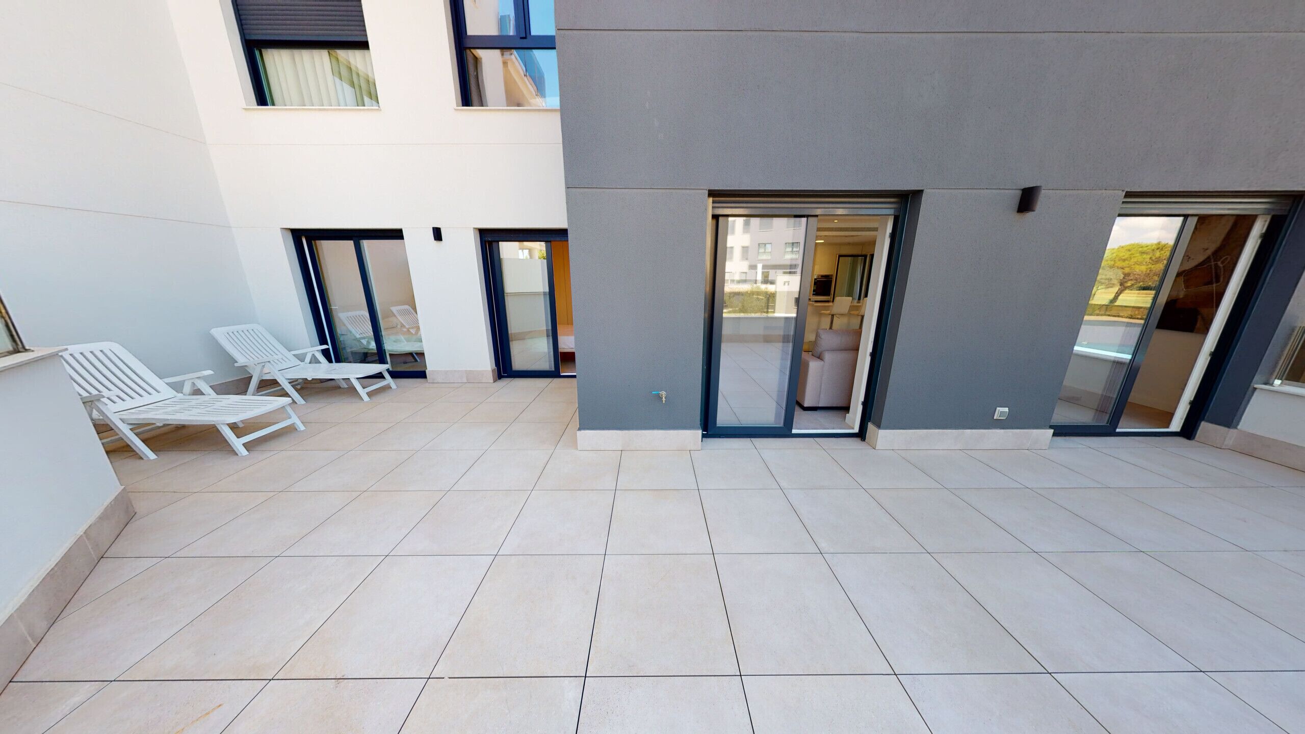 Een modern gebouw met een betegeld terras, witte lounge fauteuils en glazen deuren die leiden naar een binnenruimte.