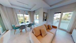 A modern living room with a beige sofa, a dining table with chairs, and large windows offering a view of the outdoors. - Veranda by ON Travel (Oliva)