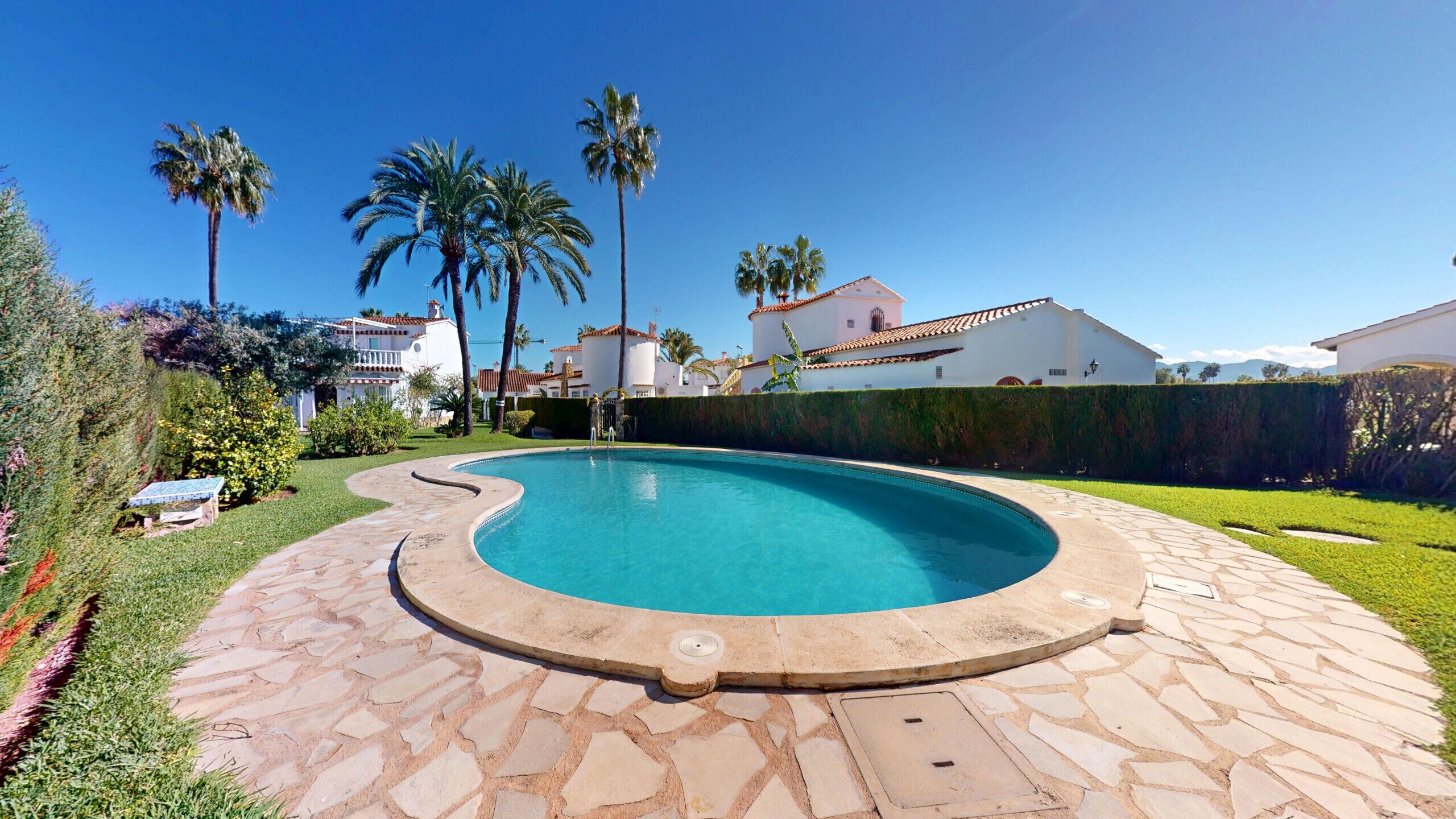 A swimming pool with a curved edge, surrounded by a stone pathway. There are palm trees and a white building with a red roof in the background.