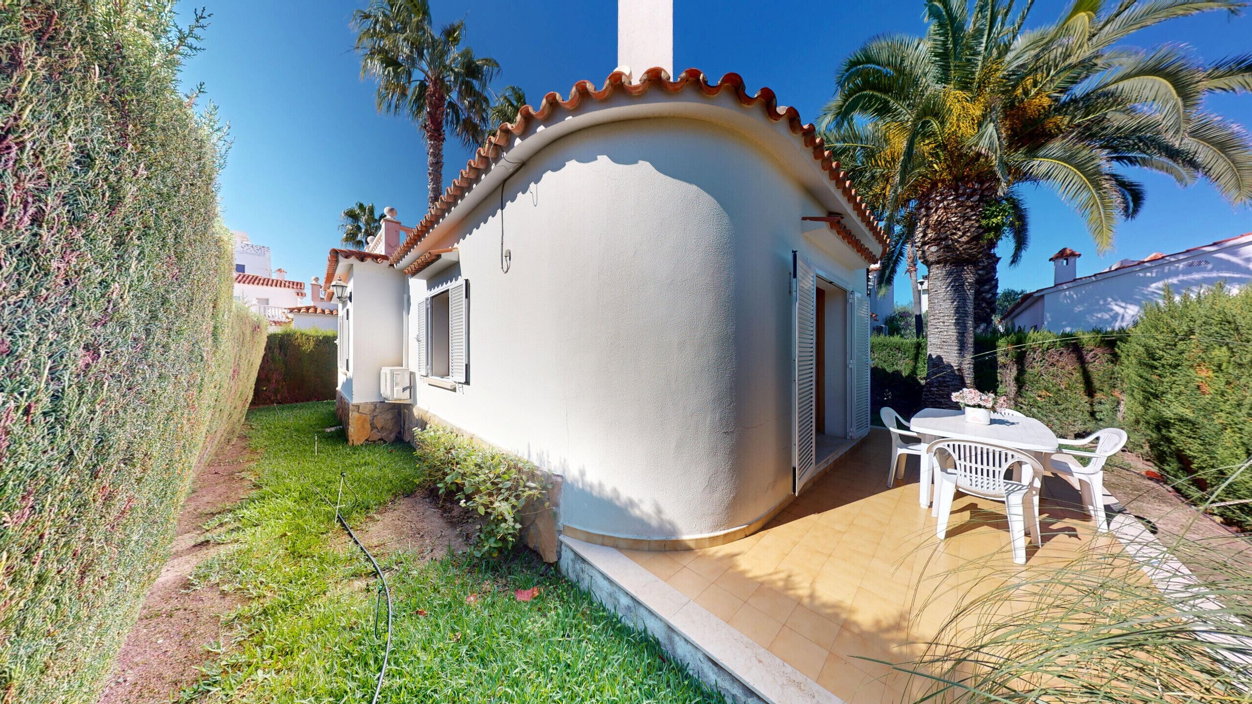 A small house with a white exterior, a tiled roof, and a patio with a table and chairs.