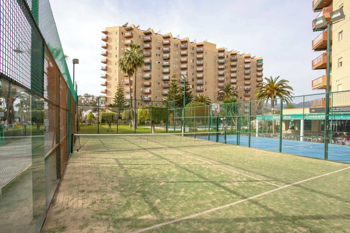 An outdoor tennis court surrounded by a fence, with a tall residential building in the background.