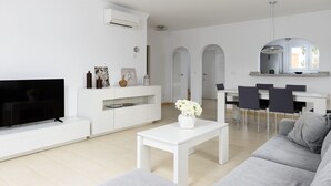 A modern living room with a flat-screen TV, white furniture, a dining area with chairs, and a kitchen visible through an archway. - Seraph by ON Travel (Oliva)