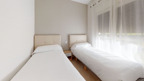 Une chambre avec deux lits simples, une table de chevet et une fenêtre avec des rideaux légers.