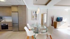 A modern kitchen with stainless steel appliances, a dining area with a glass table and white chairs, and a living space with a television and decorative plants.