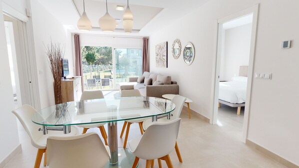 A modern living room with a glass dining table, white chairs, a sofa, and a bed in the adjacent room.
