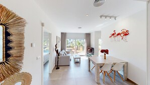 A modern living room with a sofa, coffee table, and dining area. There is a view of the outside through large windows. - Nobilis by ON Travel (Oliva)