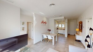 A modern living room with a flat-screen TV on a glass stand, a dining area with a white table and chairs, and a kitchen in the background.
