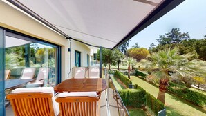 Eine moderne Terrassenanlage mit Holztisch und -stühlen, einem Glasgeländer und Blick auf einen gepflegten Garten mit verschiedenen Pflanzen und Bäumen.