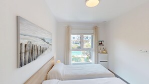 A hotel room with a bed, a painting, a window with a view of buildings, and a small cabinet with a lamp.